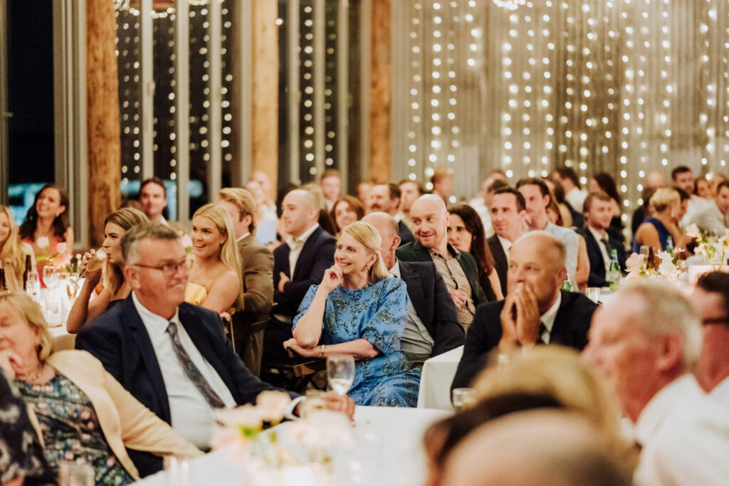 Wedding reception guests seated at long tables, smiling and listening during speeches wedding party under warm lights in a beautifully styled indoor wedding venue.
