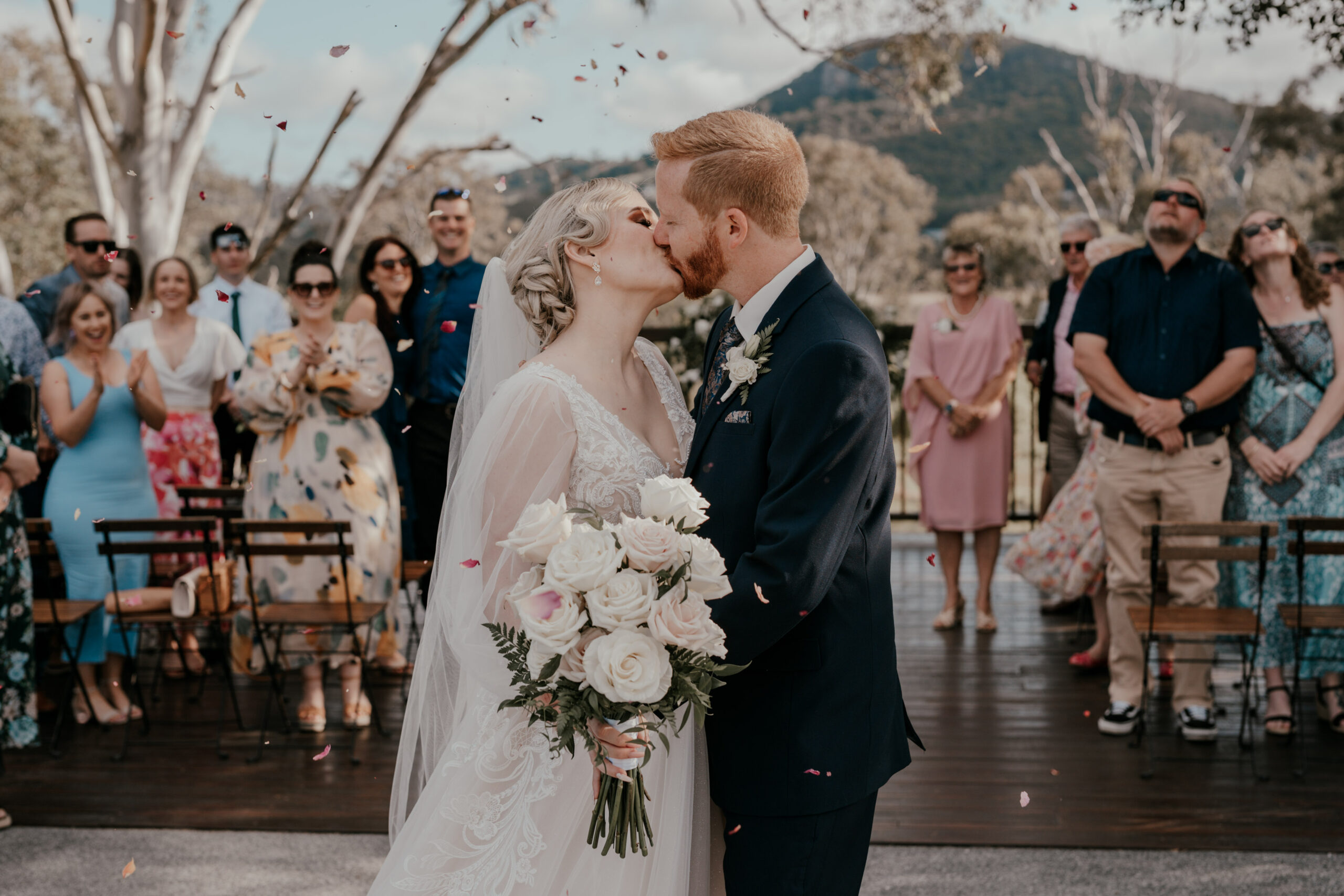 Bride and groom sharing a kiss during an outdoor country wedding venue ceremony, holding a white rose bouquet as guests celebrate behind them with rolling hills mountains in the background.