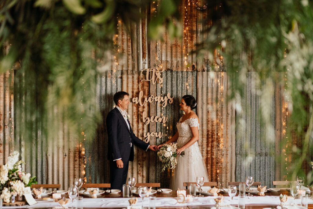 Bride and groom holding hands in front of a rustic corrugated metal backdrop wedding venue in Sunshine Coast with fairy lights and a sign reading “I’ve always been you,” set behind a beautifully styled country wedding venue reception table.