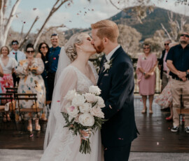 Bride and groom sharing a kiss during an outdoor wedding ceremony, holding a white rose bouquet as guests celebrate behind them with mountains in the background.